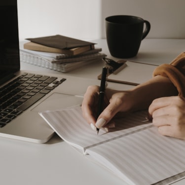 En kvinna som sitter vid ett bord och skriver i ett anteckningsblock. Framför henne står en laptop.
