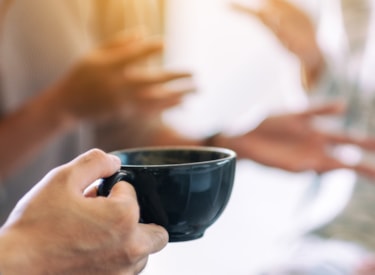 En person håller i en kaffekopp. Framför honom ser man två personer som talar med varandra. 