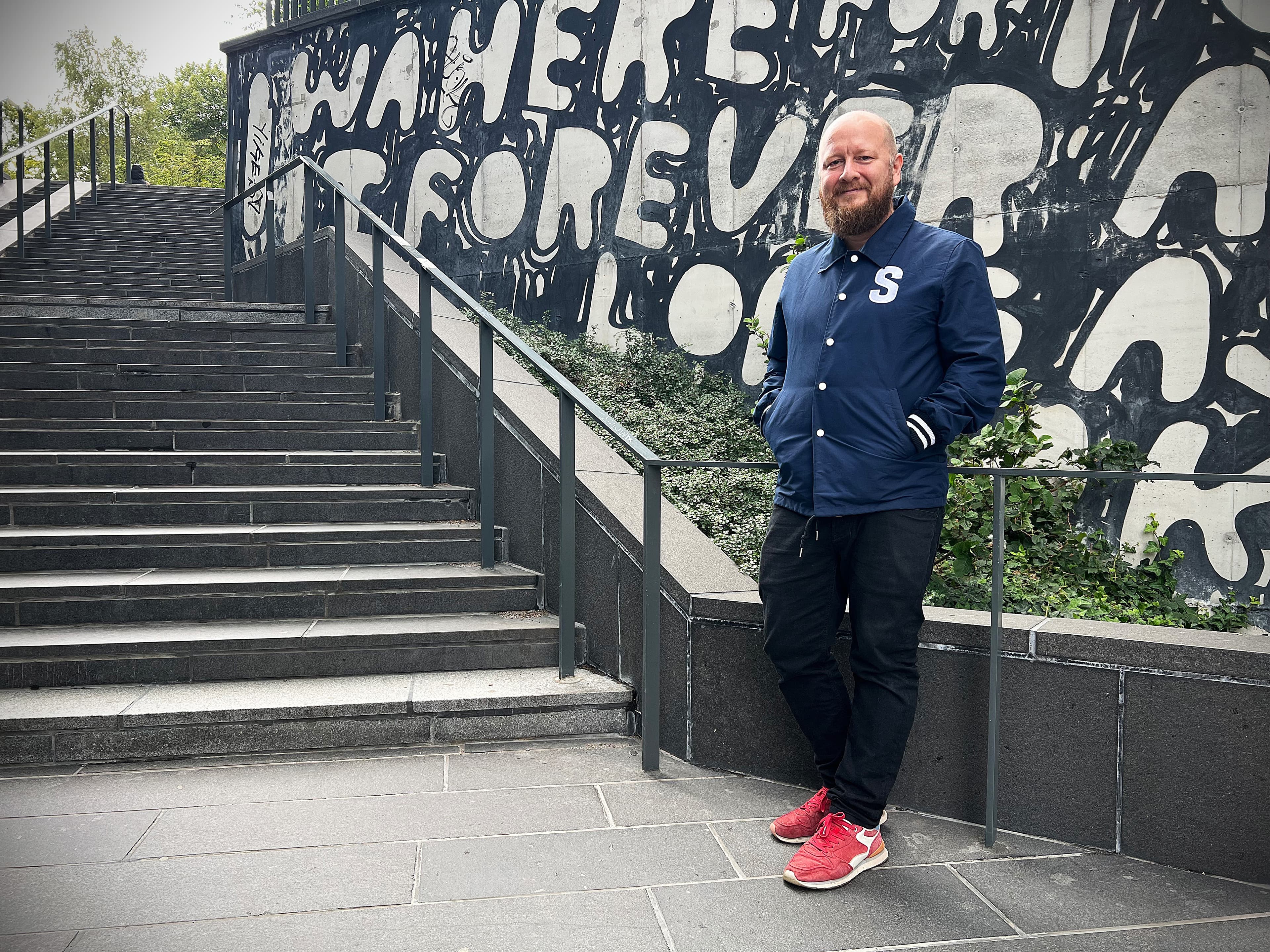 Man klädd i blå jacka, svarta byxor och röda skor. Lutandes mot en graffitivägg.