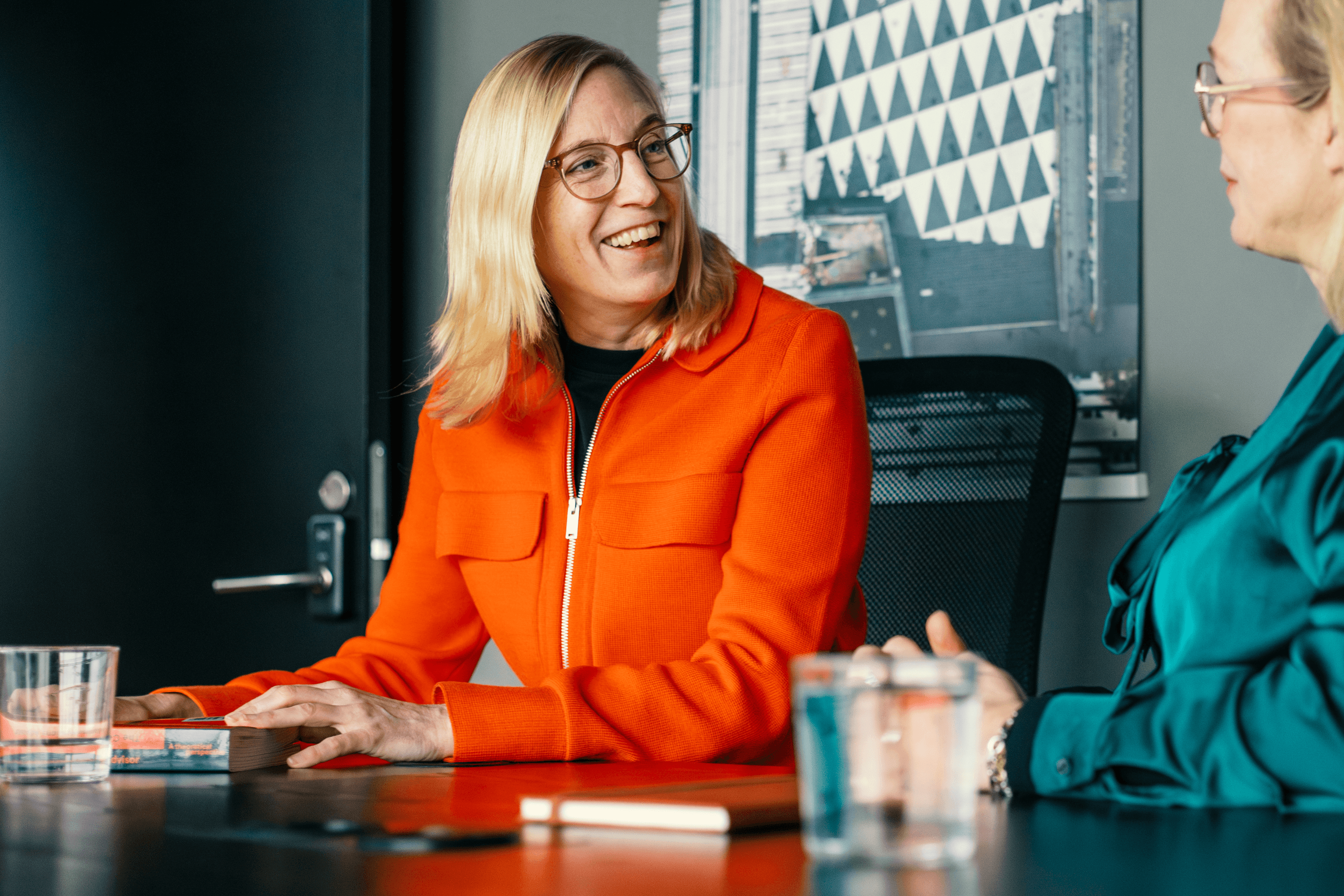 Blond kvinna i orange cardigan vid ett skrivbord som ler åt en annan kvinna.
