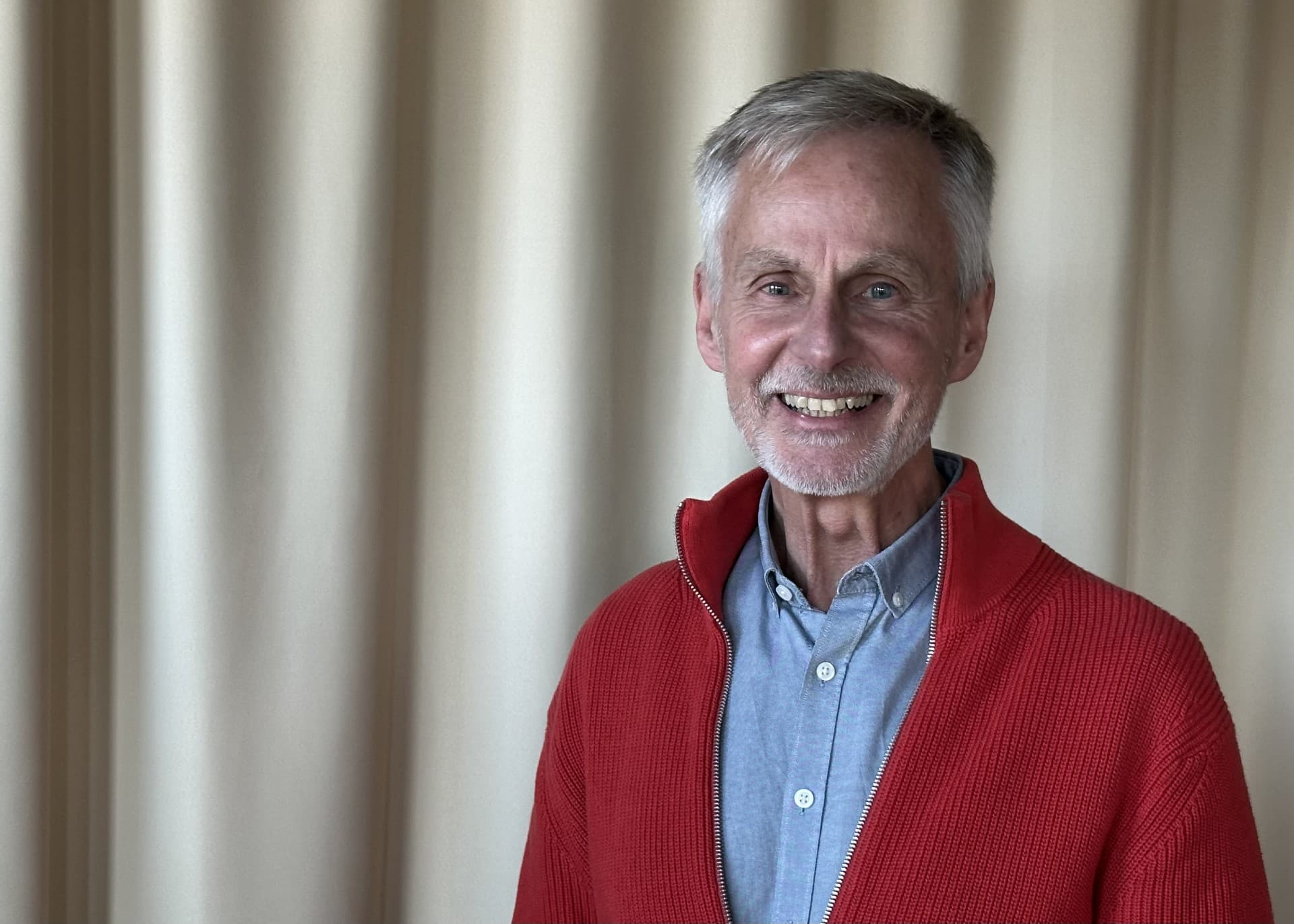 Man med grått hår, blå skjorta och röd stockad tröja ler mot kameran.