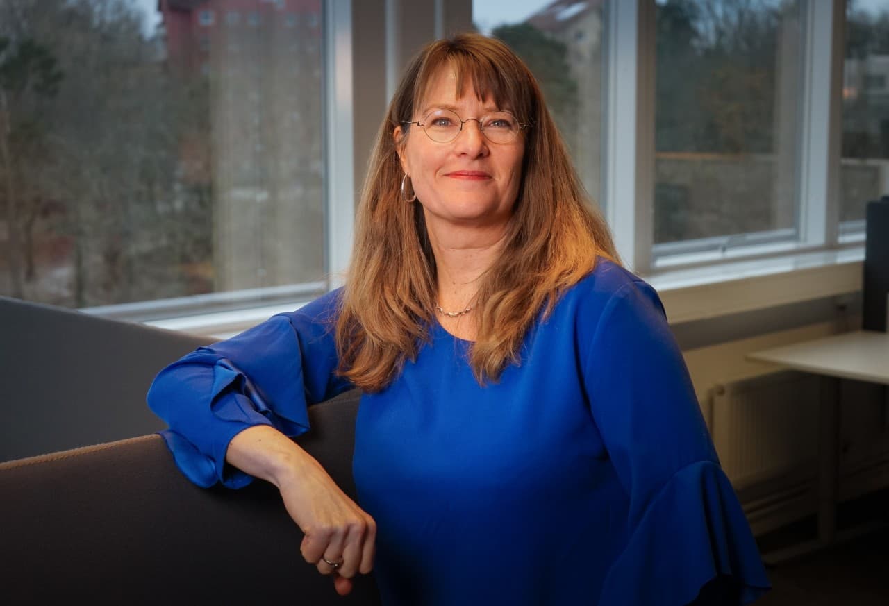 Blond långhårig kvinna i blå skjorta tittar in i kameran och lutar sig ledigt mot en fönsterkarm.