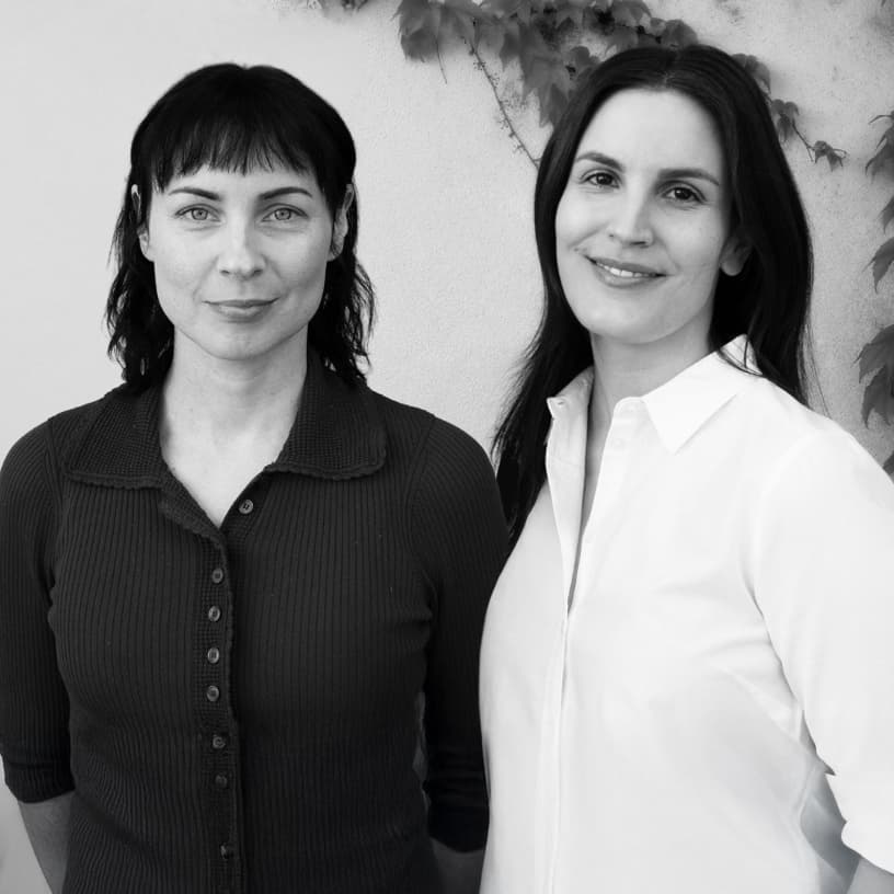 Två mörkhåriga kvinnor står och tittar leende in i kameran. 