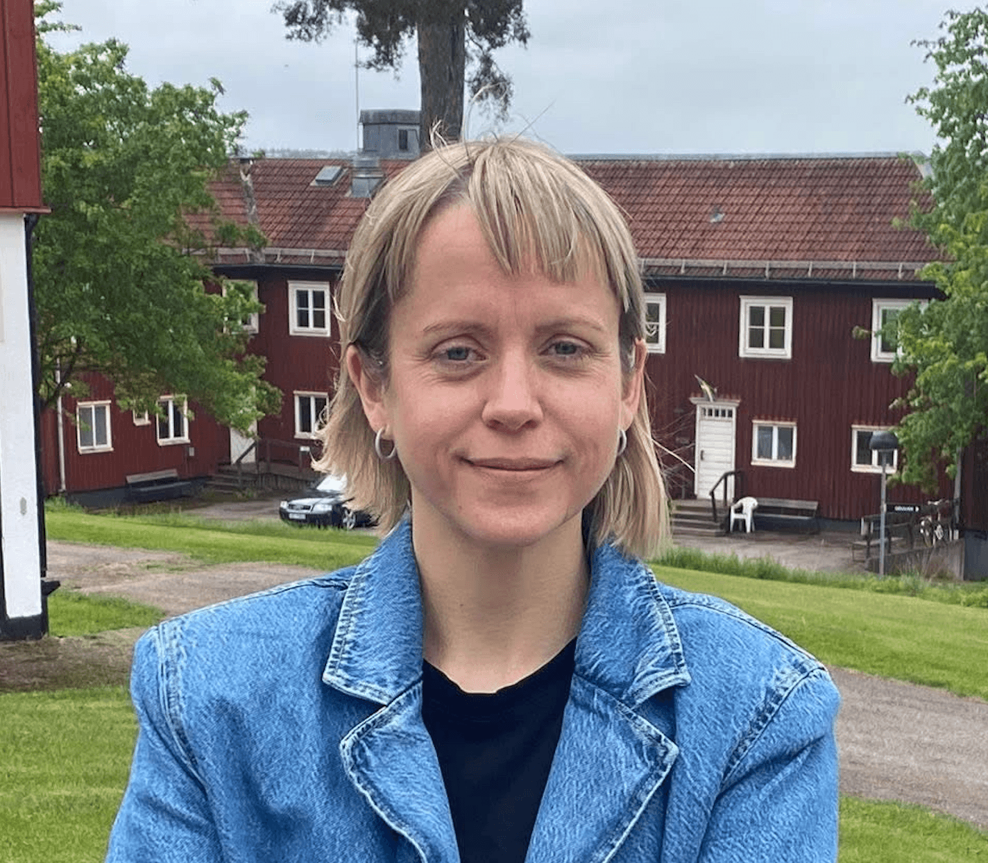 Kvinna i blå kavaj tittandes in i kameran ståendes framför ett stort rött hus