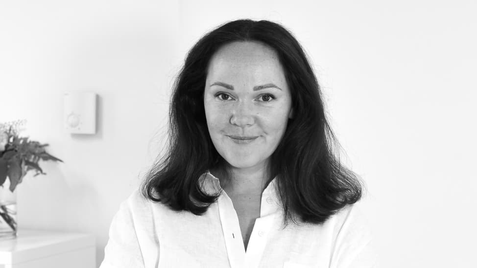 Svart/vit bild. Mörkhårig kvinna i vit blus mot en vit bakgrund tittar in i kameran