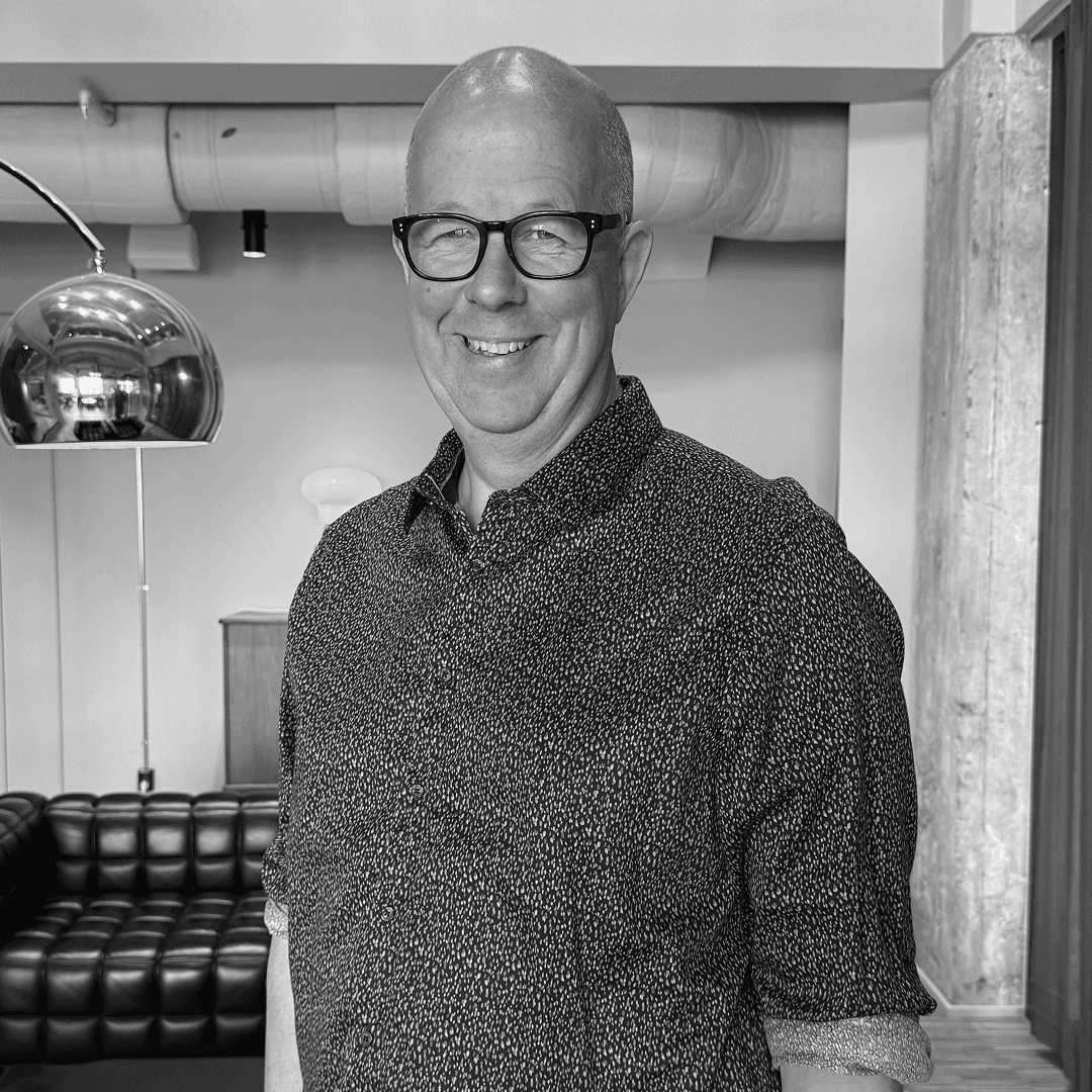 Man med inget hår, glasögon och skjorta. Ler in mot kameran.