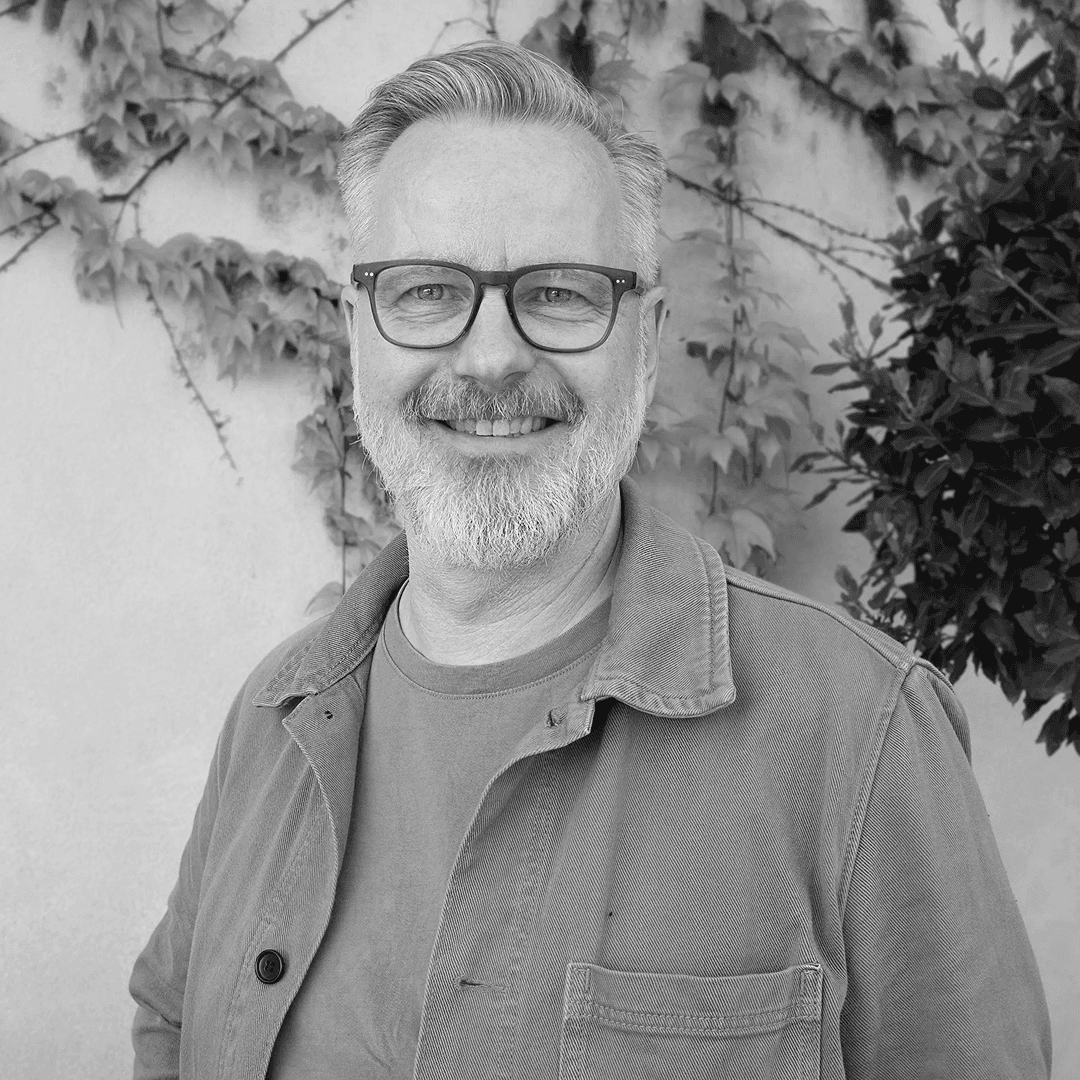 Gråhårig man med glasögon, t-shirt och skjorta ler in i kameran.