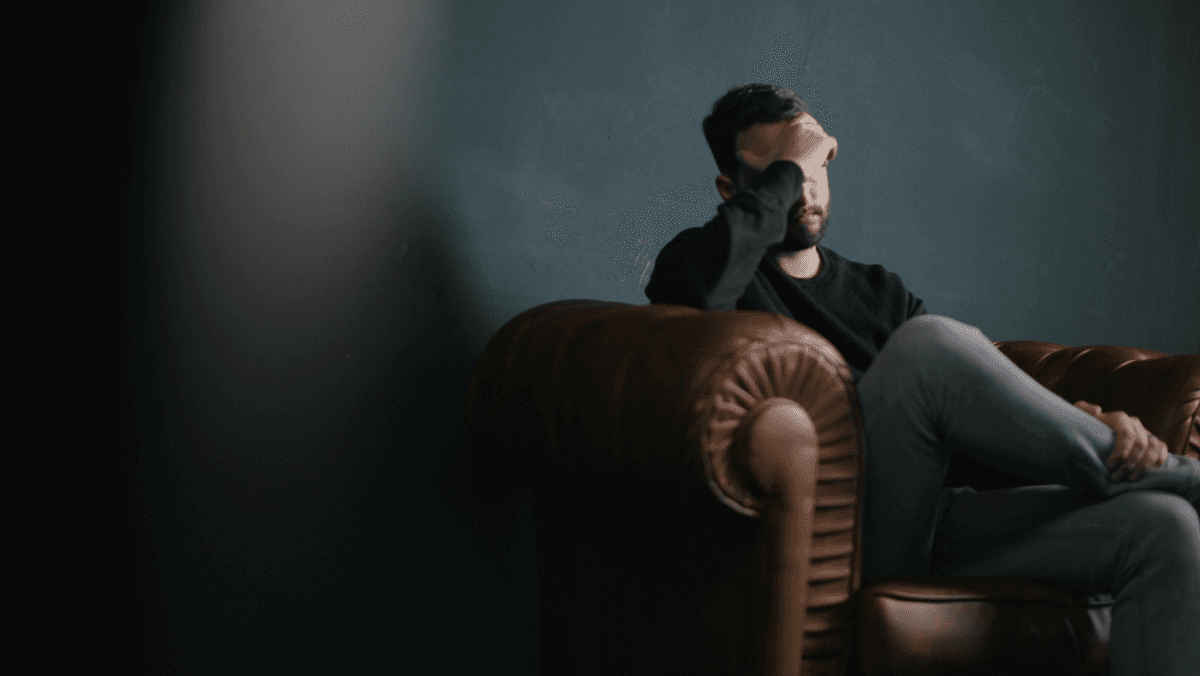 Man sitter i en soffa i en mörkt rum. Håller handen över pannan.