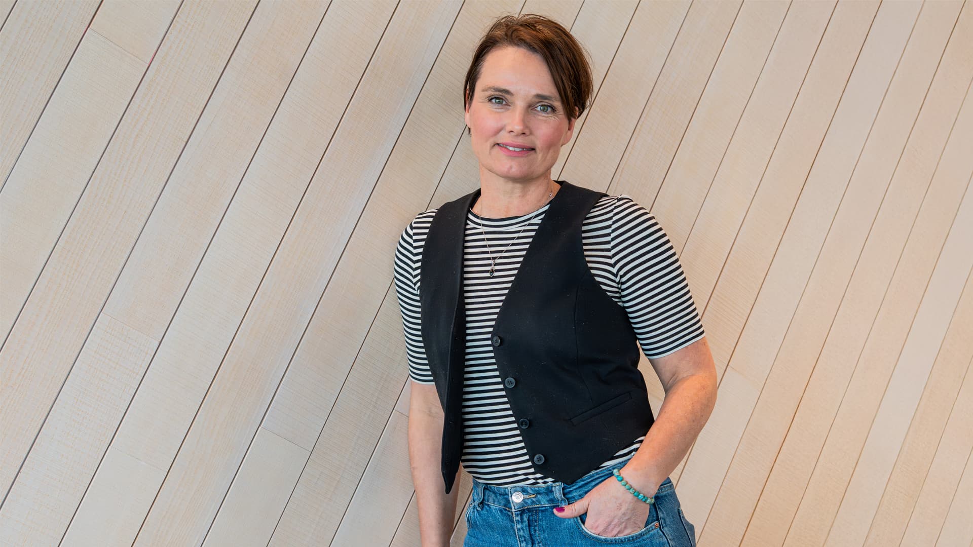 Kvinna med kortklippt brunt hår. Lutande mot en vägg med ena handen i fickan.