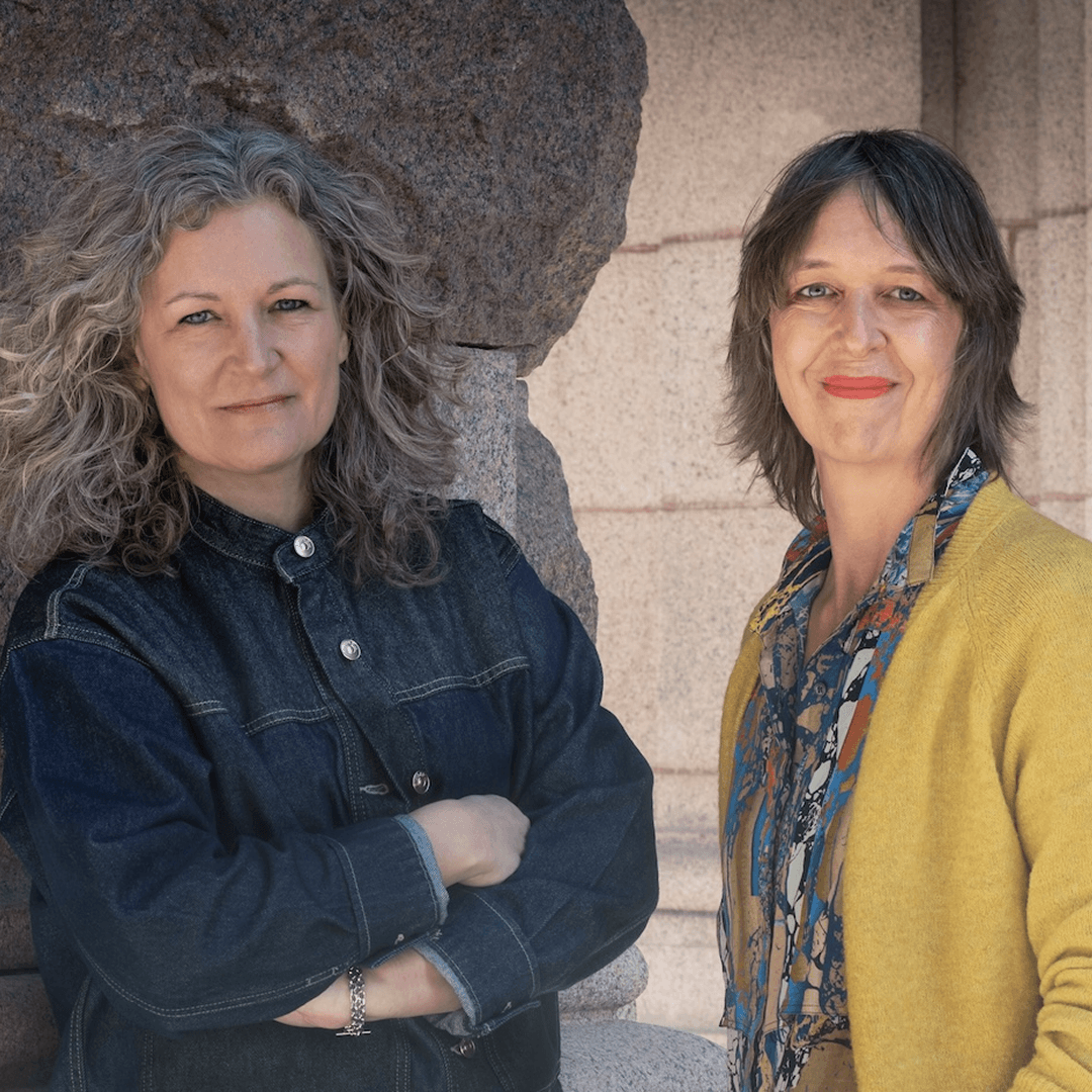 Två kvinnor. En med grått hår och en med brunt, poserar framför kameran.