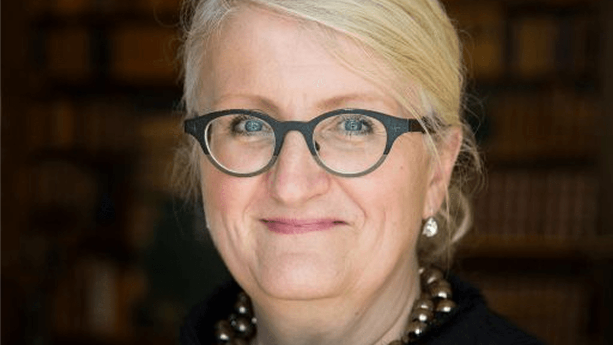 ldre, blond kvinna med svarta glasögon, ler in mot kameran.