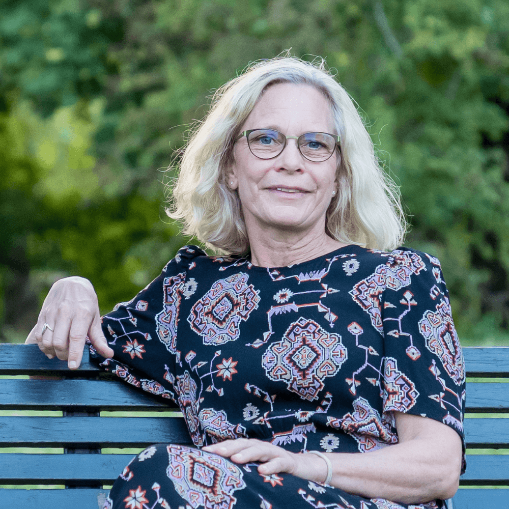 Blond kvinna sitter på en bänk och poserar framför kameran.